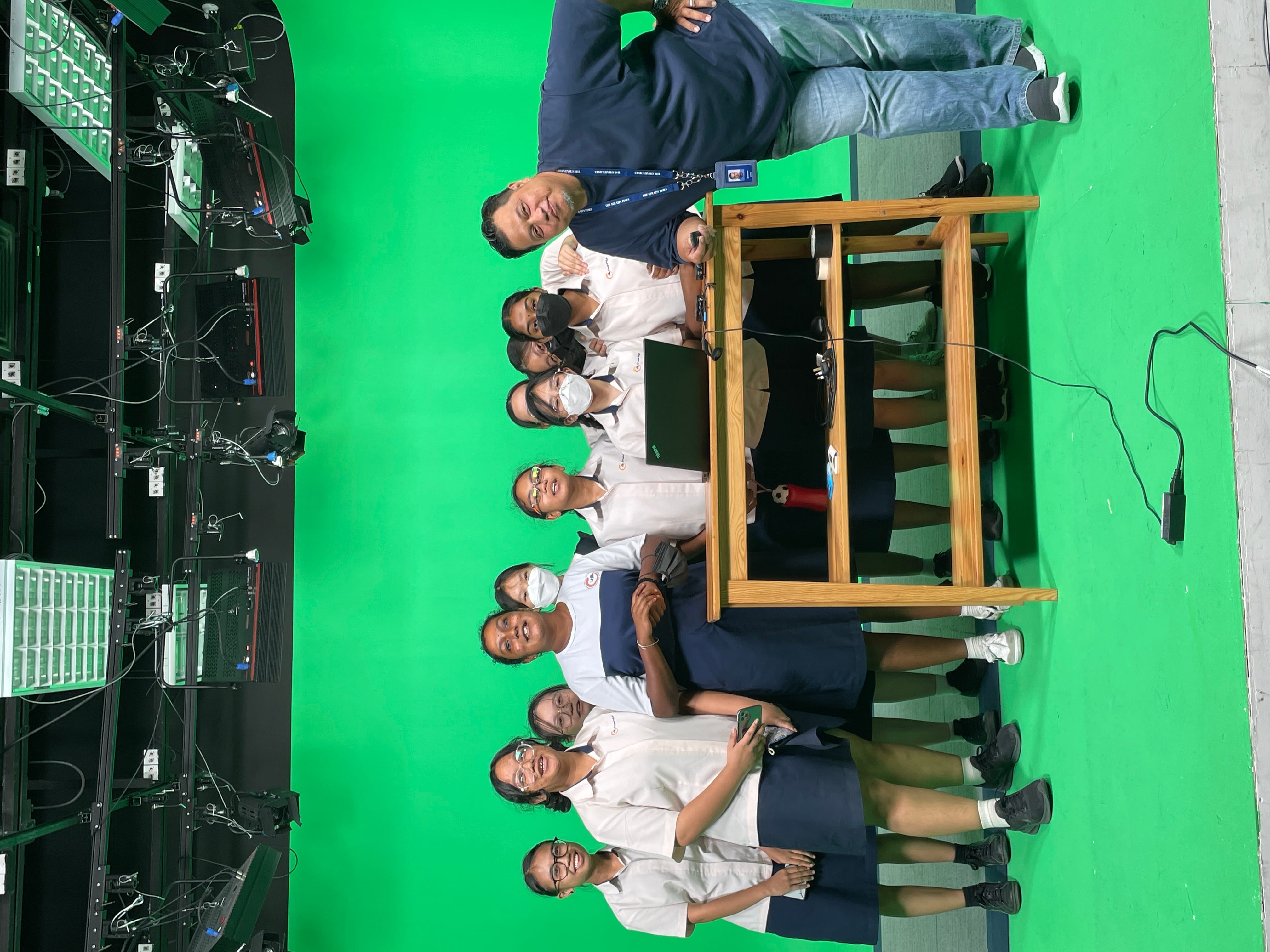 A group of uniformed students and an adult man pose together in a green screen studio with studio lights overhead.